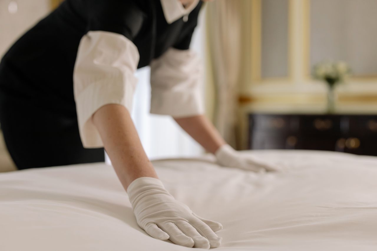 Pracovnice hotelu v bílých rukavicích uhladí čerstvě povlečenou matraci.