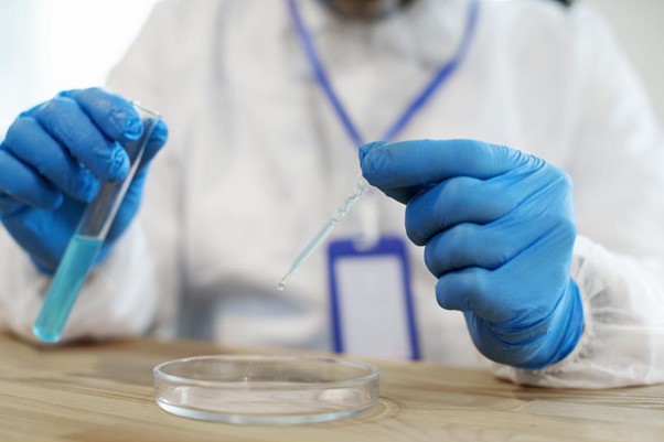 Alternativní text: Vědec v ochranném oděvu provádí experiment, při kterém pipetou nakapává tekutinu ze zkumavky do Petriho misky.