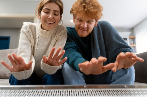 Alt text: Pár sedí pohodlně na pohovce a zahřívá si ruce u radiátoru.