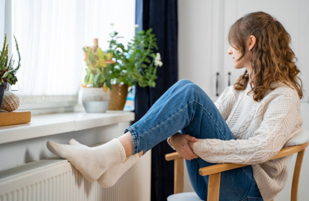Alternativní text: Žena sedí na židli a opírá si nohy o radiátor, zatímco se dívá z okna.