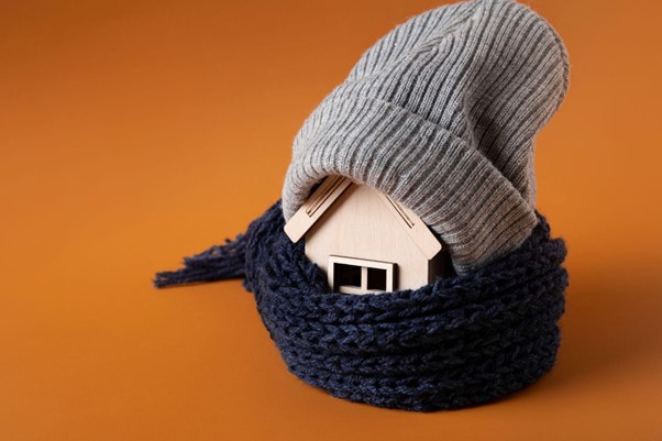 Alt-text: Malý drevený domček so šálom a čapicou ako symbol tepelnej izolácie, energetickej účinnosti a ochrany pred zimou.