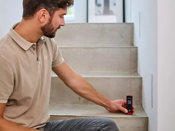 Muž měří plochu betonového schodiště pomocí laserového měřidla Einhell TC-LD 40 R; je viditelný červený laserový paprsek.