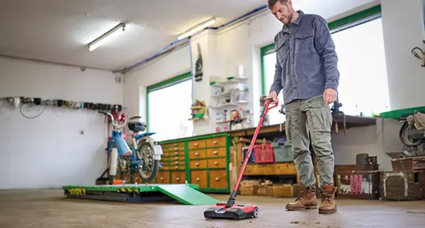Egy férfi a műhelypadlót tisztítja az Einhell TE-FS 18 Li akkumulátoros seprűjével. A műhelyben motorkerékpár-emelő és munkapadok találhatók.