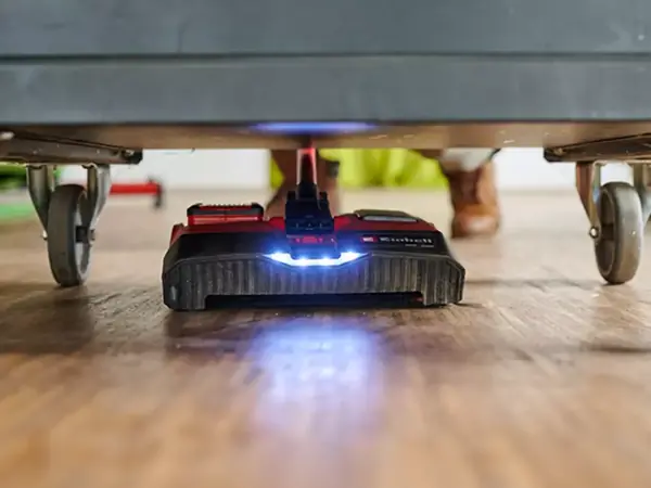 Az Einhell TE-FS 18 Li akkumulátoros seprűjének padlótisztító fején lévő LED-ek világítanak a bútor alatti terület takarítása közben.
