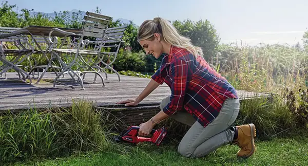 Žena klečí na zahradě a stříhá trávu na okraji terasy pomocí aku nůžek na trávu a keře Einhell GE-CG 18/100 Li-Solo.