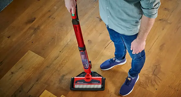 Osoba čistí dřevěnou podlahu akumulátorovým čističem tvrdých podlah Einhell CLEANEXXO; čisticí válec je umístěn v čisticí stanici.