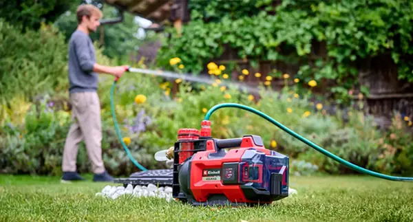 Ein Mann bewässert den Garten mit der Einhell Akku-Gartenpumpe AQUINNA 18/30 F LED AUTOMATIC, die im Vordergrund auf dem Rasen steht.