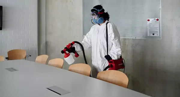 Eine Person in Schutzkleidung nutzt die Einhell Farbsprühpistole TC-SY 500 P zur Desinfektion von Stühlen in einem Besprechungsraum.