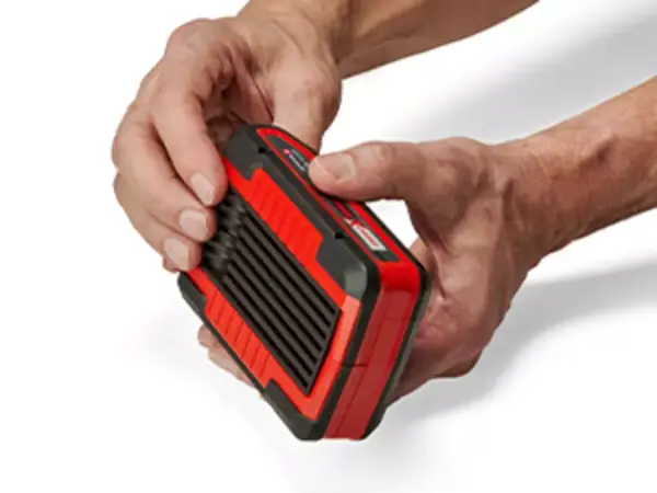 Dvě ruce drží akumulátor Einhell Power X-Change za vroubkovanou prohlubeň; viditelné je robustní pogumované pouzdro.