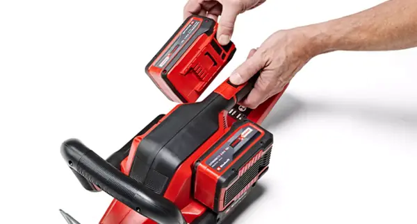 Akumulátor Einhell Power X-Change je vkládán rukou do zahradního nářadí se dvěma sloty na akumulátory, na bílém pozadí.