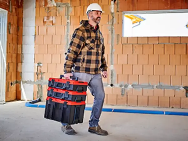 Muž nese několik stohovaných kufrů Einhell E-Case L with wheels přes staveniště.