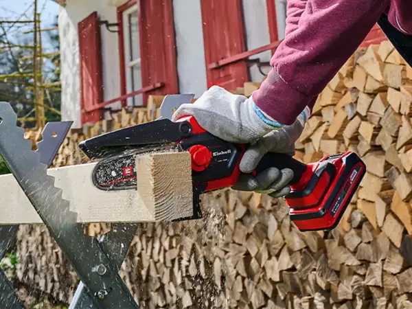 Žena obouruč vede akumulátorovou prořezávací pilu při řezání kusu dřeva před hromadou dřeva.