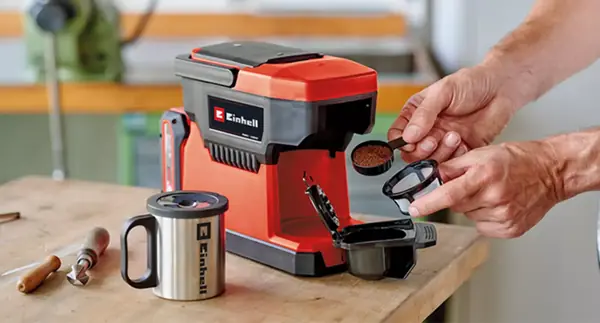 Eine Person füllt Kaffeepulver in den Filter der Einhell Akku-Kaffeemaschine TE-CF 18 Li, daneben steht ein Edelstahlbecher.